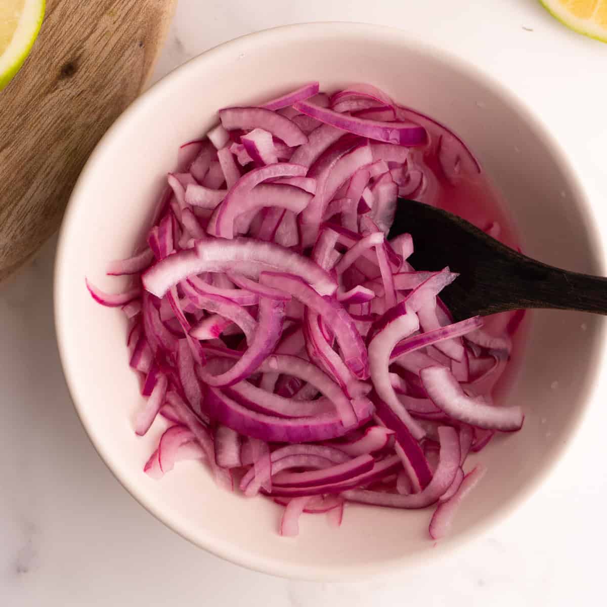 Lime pickled onions in a white bowl with a wooden spoon.