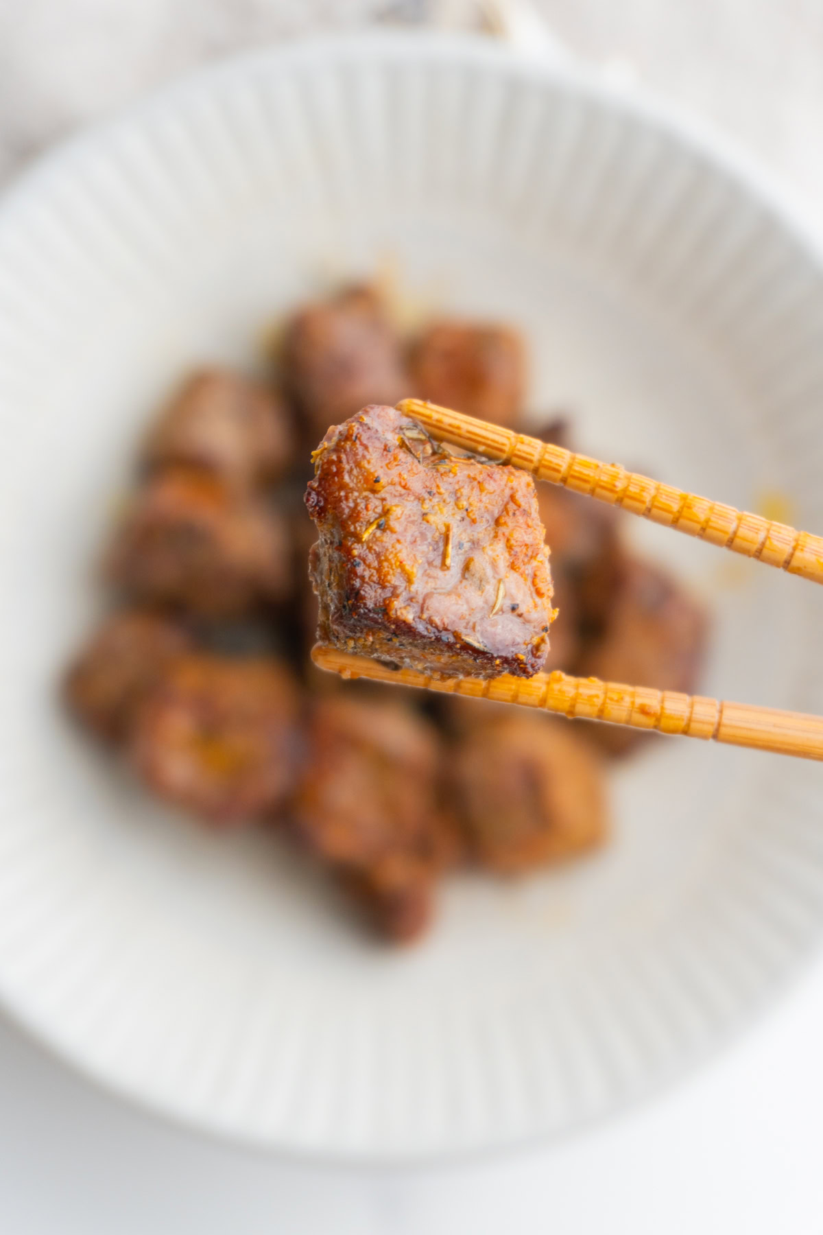 Close up shot of a steak bite in chopsticks with more steak bites in the background.