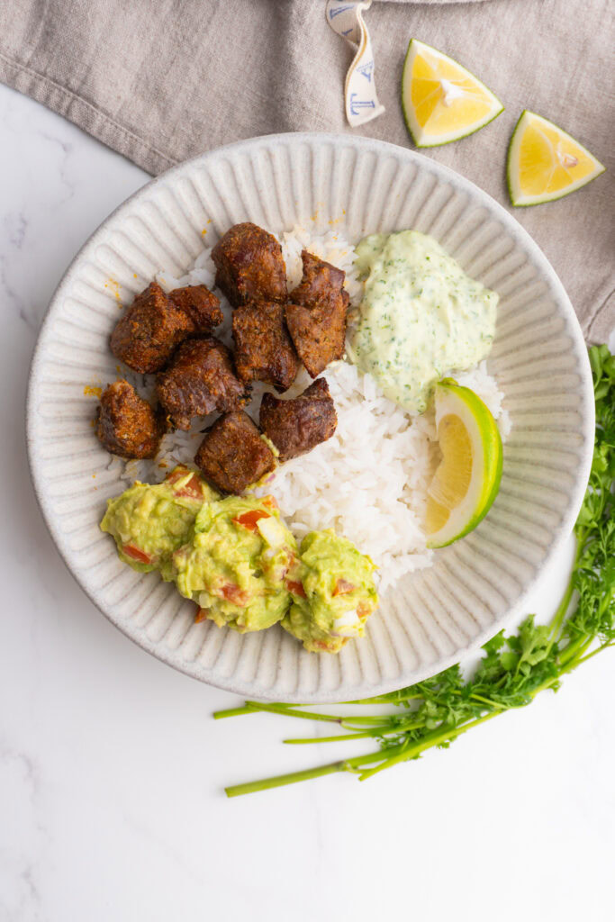 Bowl served with rice, steak bites, cilantro sauce, guacamole and lime wedges.