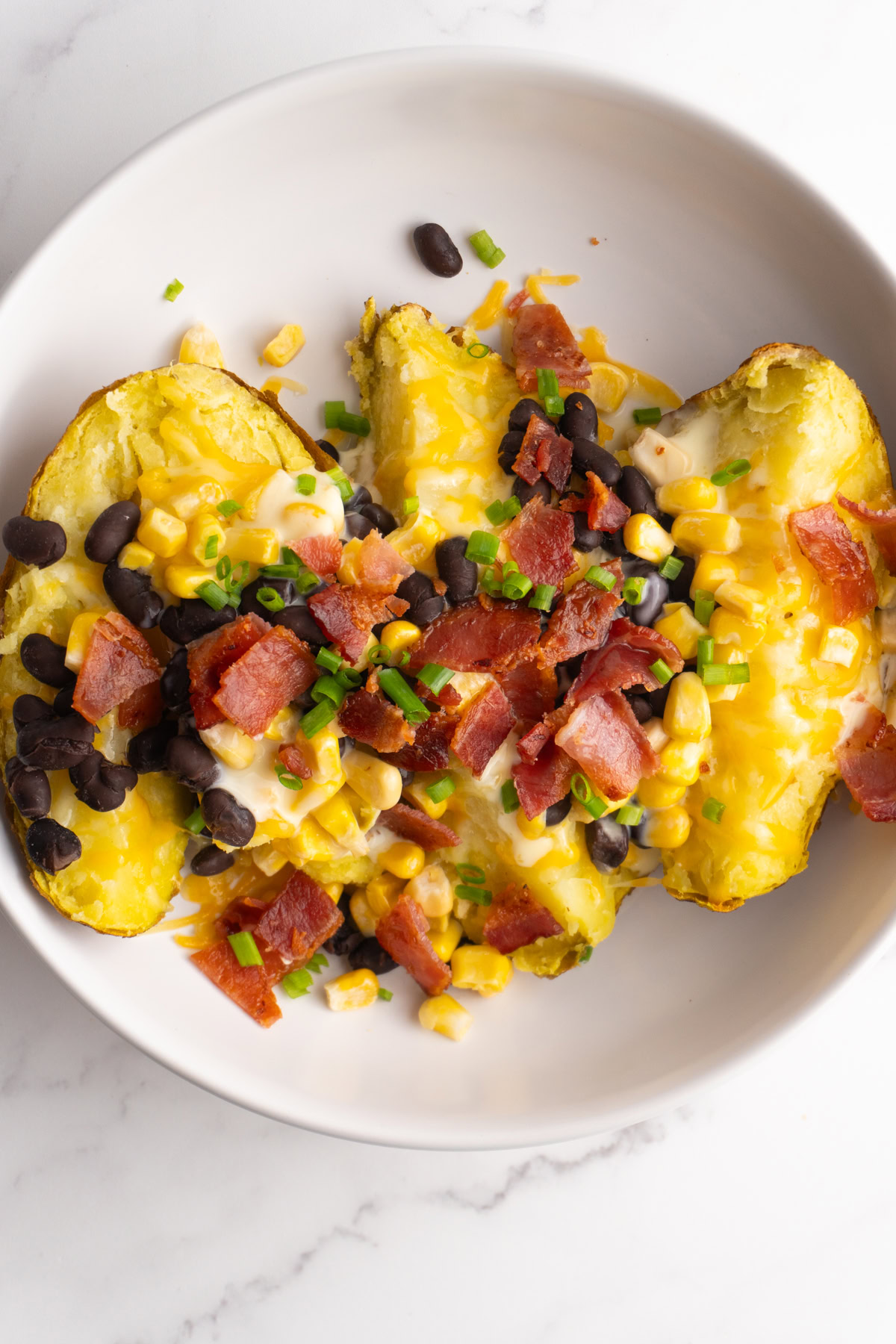Baked potato broken open in a bowl topped with black beans, corn, bacon, and green onions.