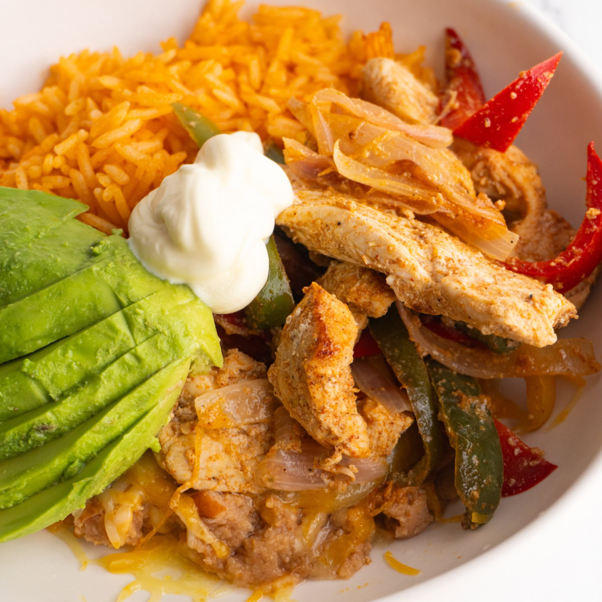 Chicken fajitas served in a bowl with rice, refried beans, avocado and sour cream.