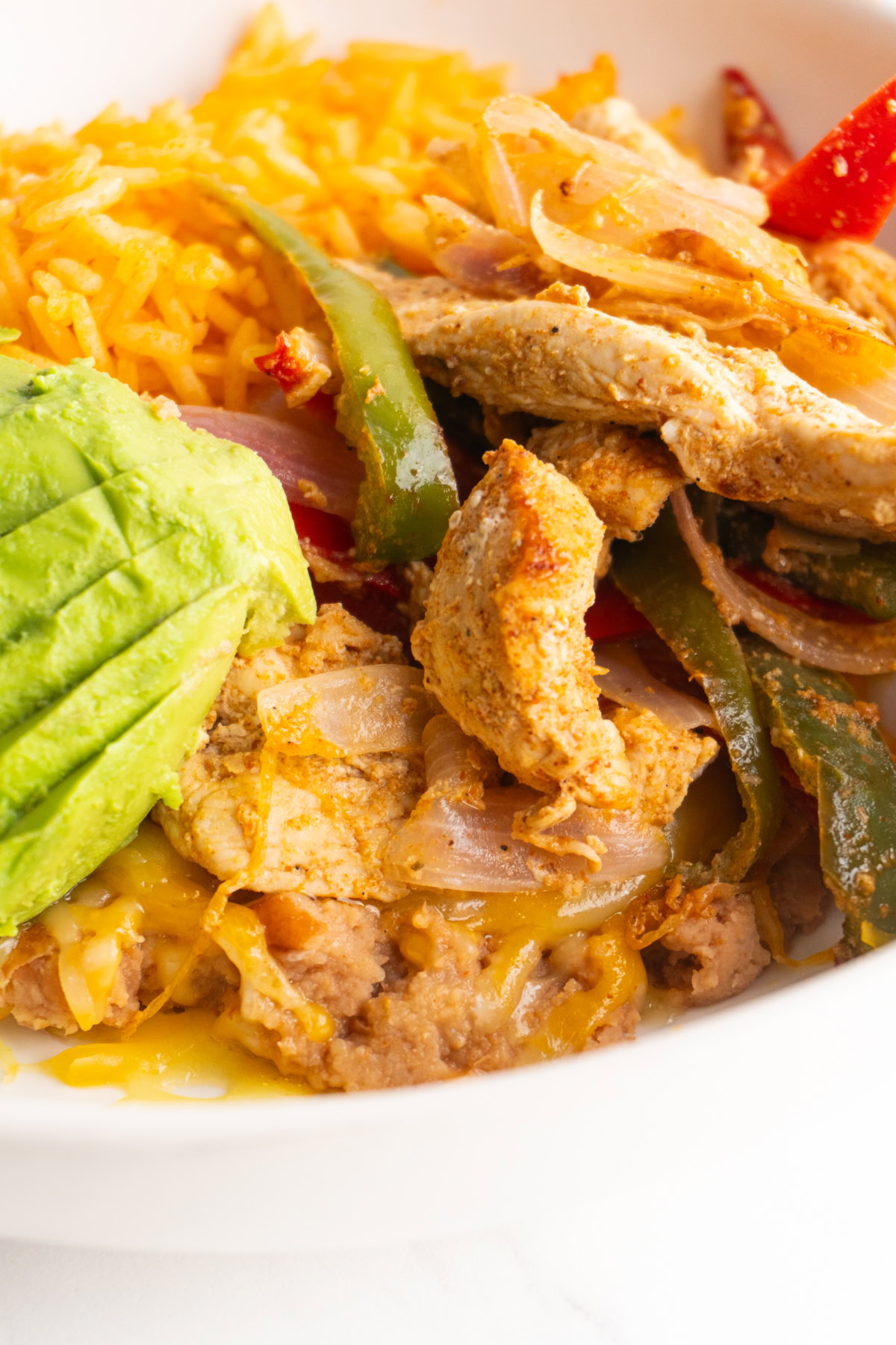 Side view of a chicken fajita bowl with refried beans, cheese, rice, chicken fajitas and avocado.
