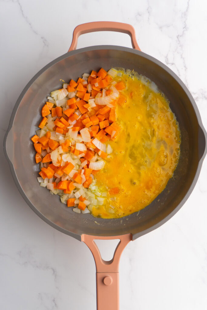 Scrambled eggs added to one half of the skillet next to cooked carrots and onions.