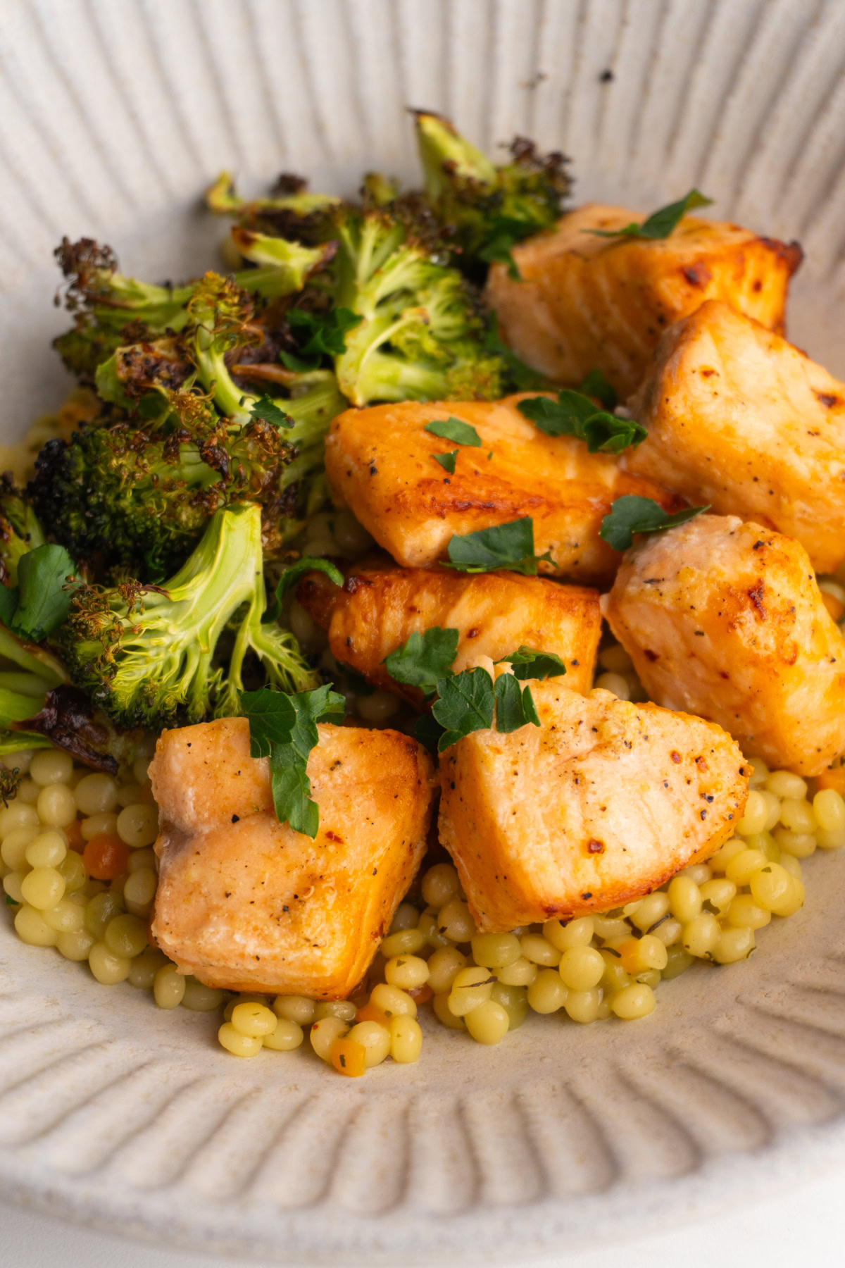 Side view of salmon bites on bed of couscous with broccoli in a bowl.