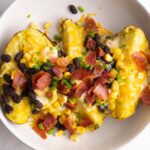Loaded baked potato served in a white bowl topped with cheese, beans, bacon and corn.