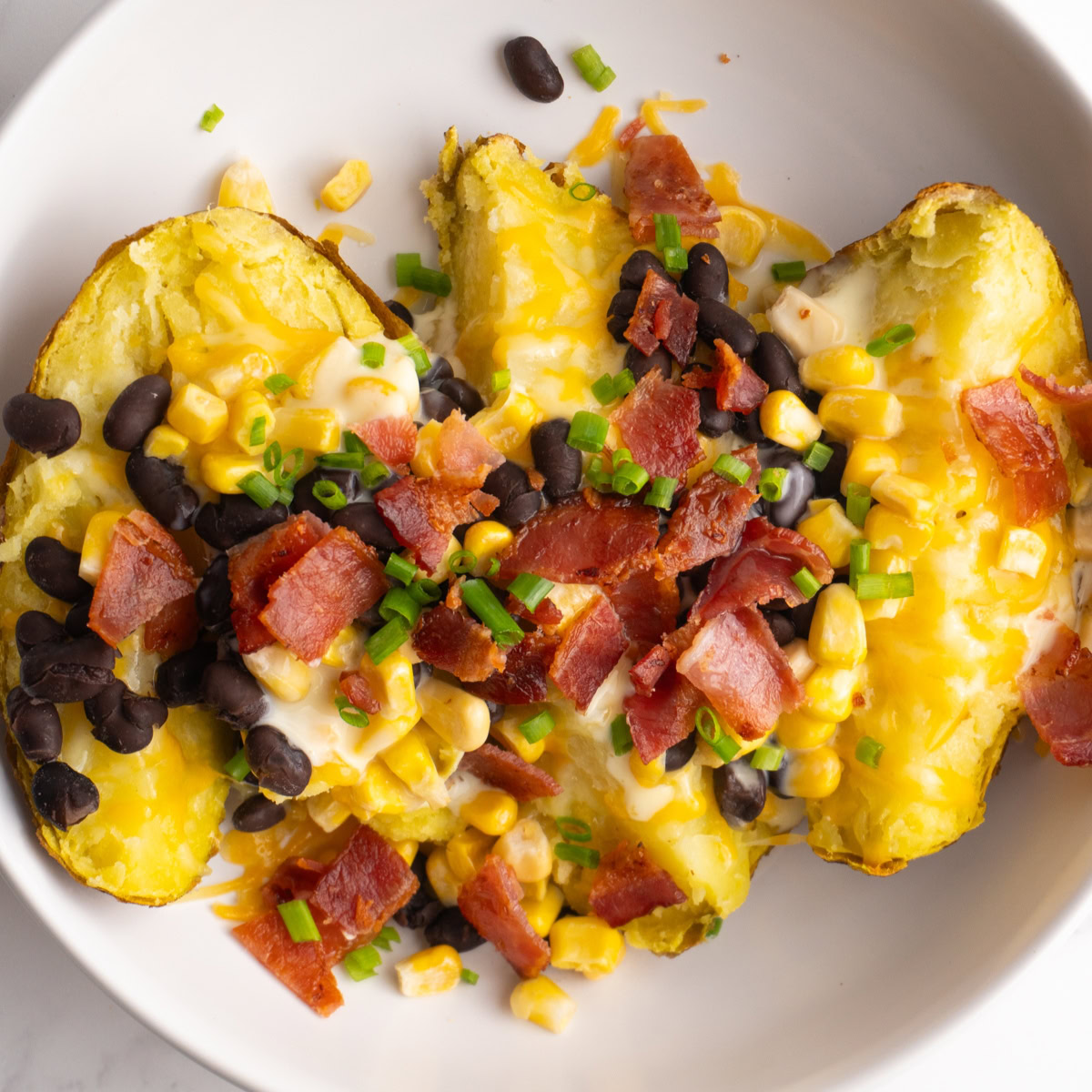 Loaded baked potato served in a white bowl topped with cheese, beans, bacon and corn.