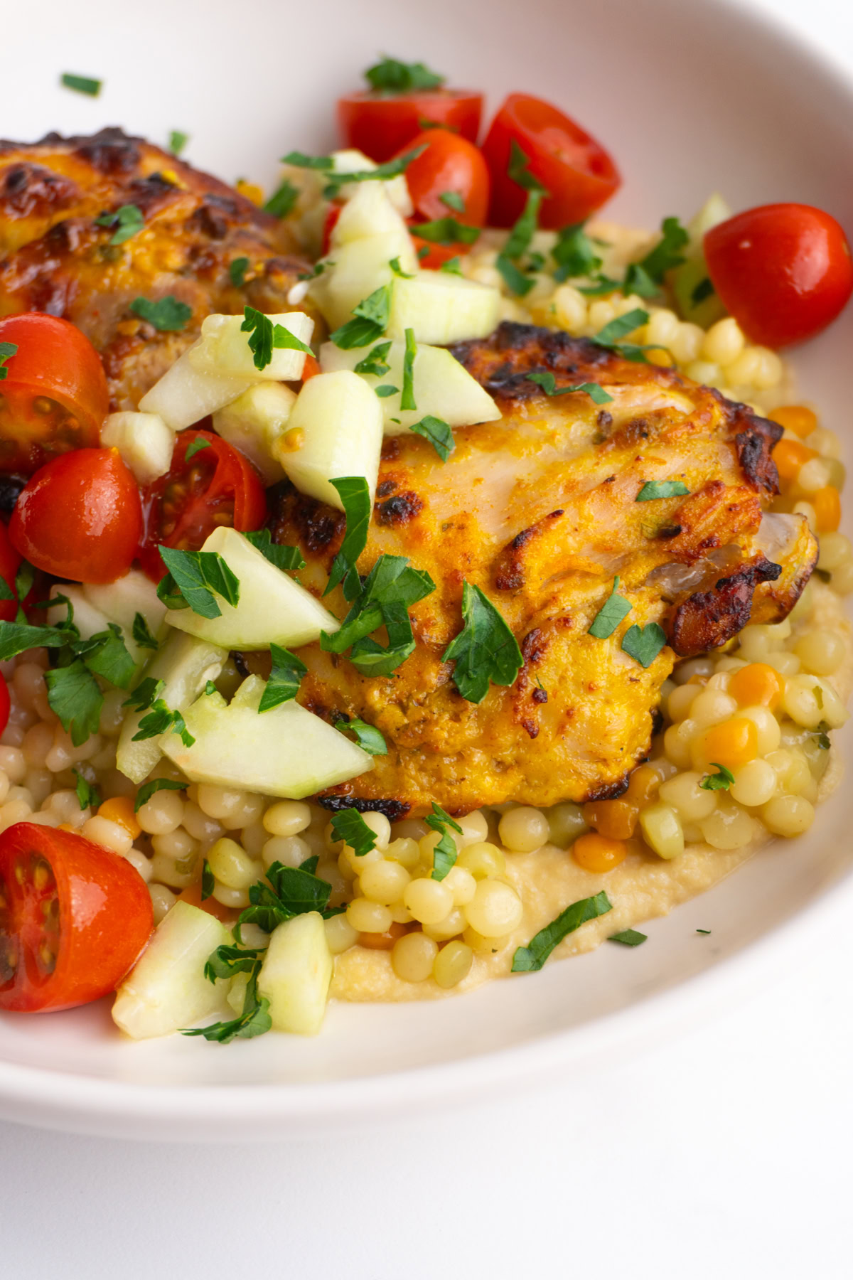Side view of chicken thigh on a bed of hummus and couscous topped with cucumbers and tomatoes.