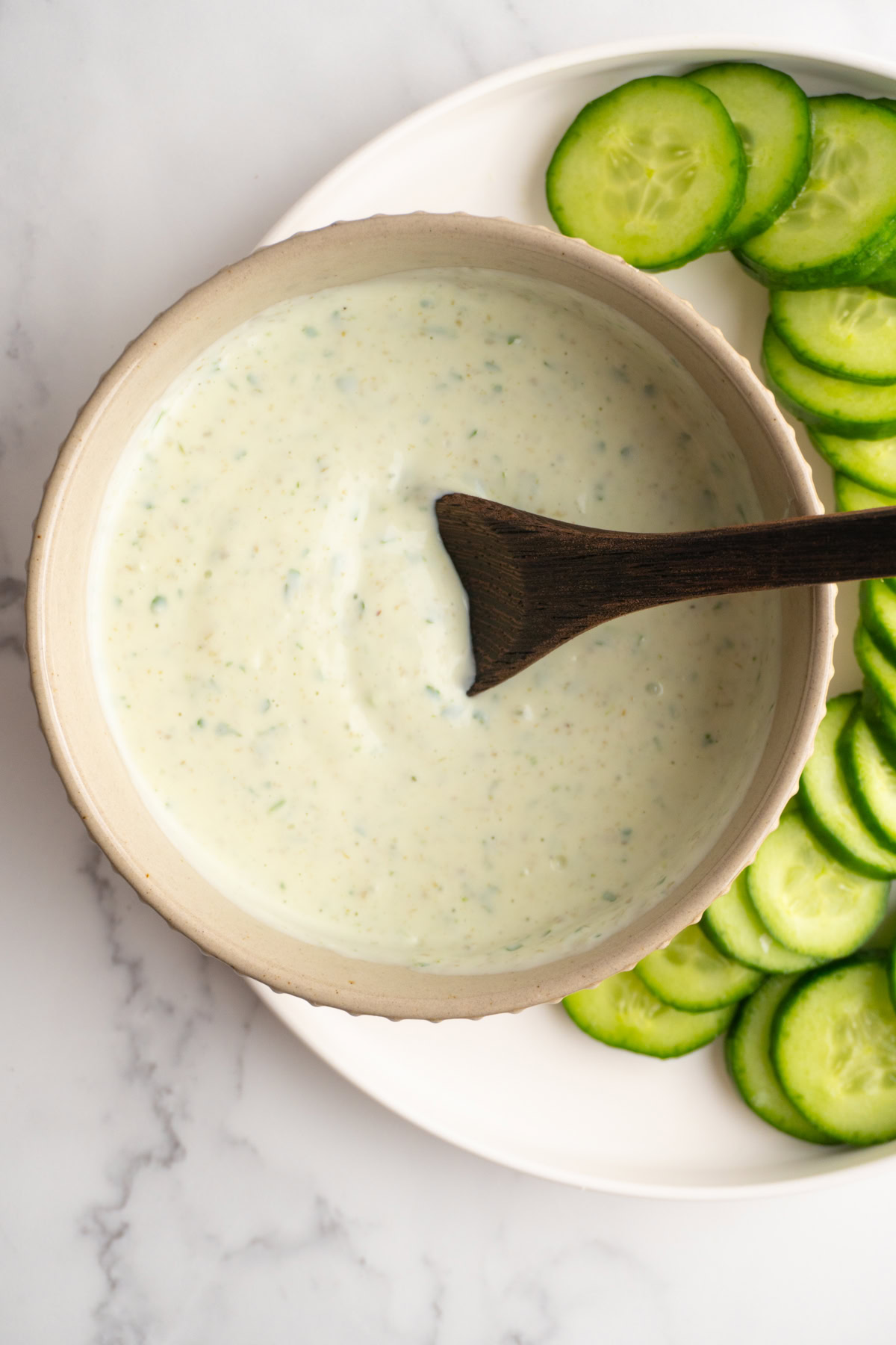 Green goddess dip served in a small bowl with a wooden spoon next to sliced cucumbers.