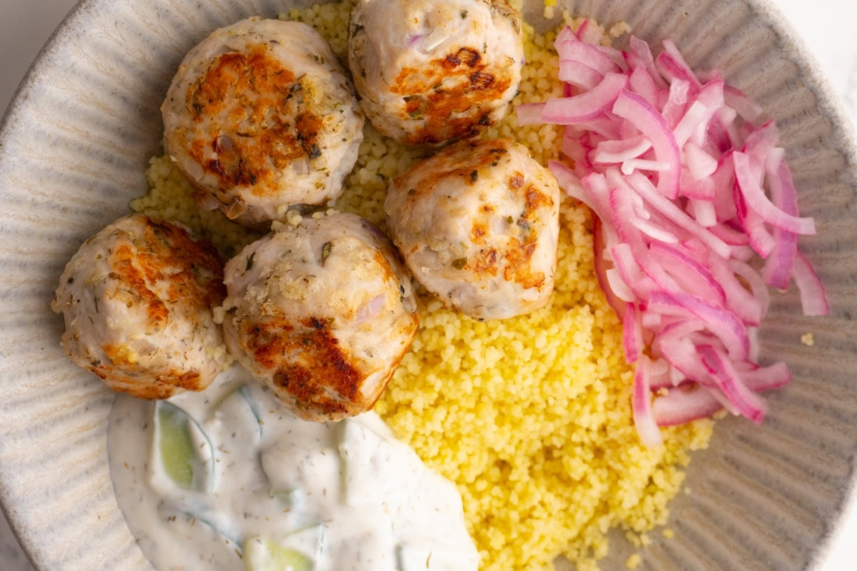 Close up of Greek turkey meatballs on a bed of couscous with sides of tzatziki and red onions.