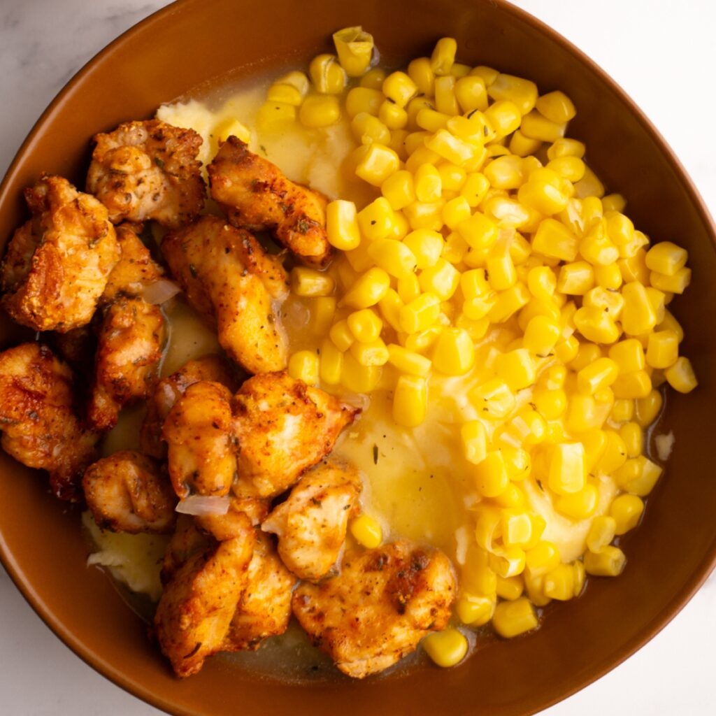 Brown bowl served with mashed potatoes, corn and chicken bites with gravy on top.