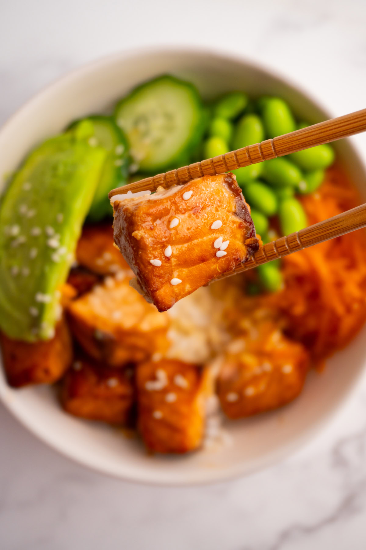 Close up of a piece of salmon being held by chopsticks with a poke bowl underneath it.