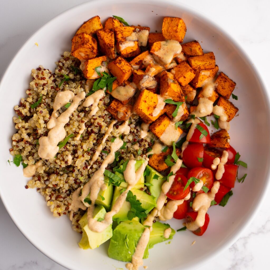 White bowl served with roasted sweet potatoes, quinoa, veggies, and taco sauce.