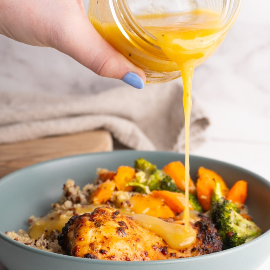 Hand pouring honey dijon vinaigrette over a bowl of chicken and veggies.